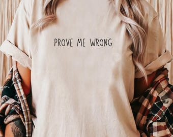 May include: Beige t-shirt with the phrase "PROVE ME WRONG" printed in black. The shirt is worn with a plaid flannel shirt. The sleeves of the t-shirt are rolled up.