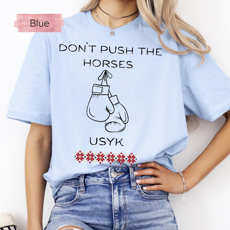 May include: Light blue t-shirt with the text "DON'T PUSH THE HORSES" and "USYK" in black. The shirt features a graphic of two boxing gloves. A red and white floral design is at the bottom of the shirt. The word "Blue" is in a pink rectangle.