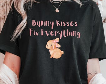 May include: Black t-shirt with the text "Bunny Kisses Fix Everything" in pink script. A watercolor illustration of a brown bunny with a pink bow is below the text. The shirt is a casual, everyday wear item.