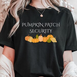 May include: Black t-shirt with the text "PUMPKIN PATCH SECURITY" in white. Below the text is a row of illustrated pumpkins in various shades of orange, yellow, and green. The shirt is a casual style.