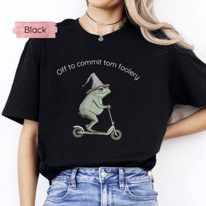 May include: Black t-shirt with a humorous design. It shows a green frog in a wizard hat on a scooter, with the text "Off to commit tom foolery" above. A pink rectangle with the word "Black" is in the upper left.