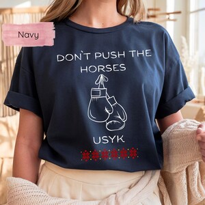 May include: Navy blue t-shirt with the text "DON'T PUSH THE HORSES" and "USYK" in white. The design includes a pair of boxing gloves. A red and gold floral pattern is at the bottom of the shirt.