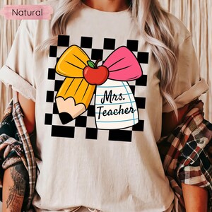 Puede incluir: Camiseta beige con un dise&ntilde;o para profesores. El dise&ntilde;o incluye un l&aacute;piz amarillo, un lazo rosa y una manzana roja, sobre un fondo a cuadros blanco y negro. Las palabras "Mrs. Teacher" est&aacute;n escritas en un bloc de notas.