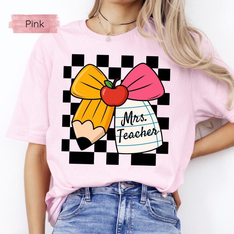 Puede incluir: Una camiseta rosa claro con un dise&ntilde;o para profesores. El dise&ntilde;o incluye un l&aacute;piz amarillo, un lazo rosa y una manzana roja, sobre un fondo a cuadros blanco y negro. Las palabras "Mrs. Teacher" est&aacute;n escritas en un bloc de notas.