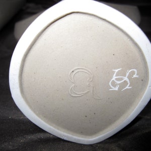 Peut inclure: Gros plan du fond d'un vase en c&eacute;ramique blanche avec une marque du fabricant qui se lit "G & S" en cursive.