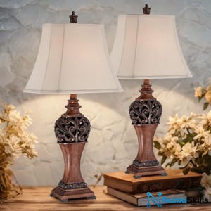 May include: A pair of table lamps featuring off-white fabric shades and dark brown, carved resin bases with intricate scrollwork details. The lamps stand on a wooden surface, each topped with a small, dark brown finial at the peak of the shade.