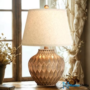 May include: A table lamp featuring a textured, bronze-toned, diamond-patterned base and a tapered, off-white fabric lampshade. The lamp is topped with a small, spherical finial and emits a soft, warm glow from within the shade.