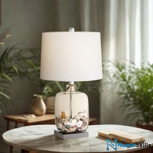 May include: A table lamp featuring a cylindrical white fabric lampshade and a clear glass base filled with a collection of assorted seashells. The lamp has a silver-toned metal neck and sits on a square base, providing a clear view of the shells.