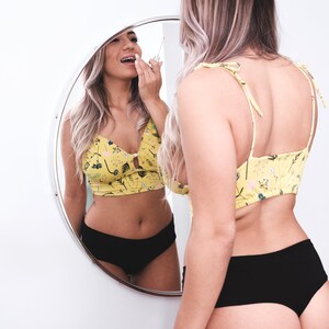 May include: A woman wearing a yellow floral crop top and black underwear is applying lip gloss while looking in a round mirror.
