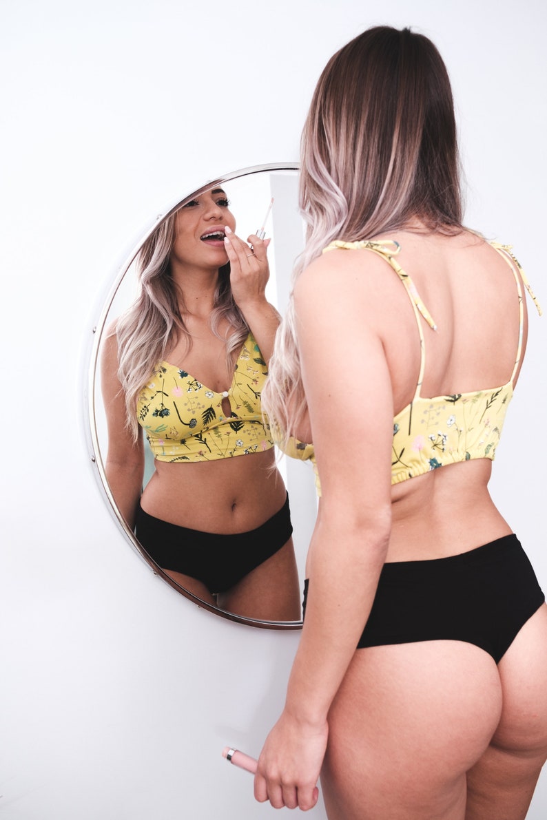 May include: A woman wearing a yellow floral crop top and black underwear is applying lip gloss while looking in a round mirror.