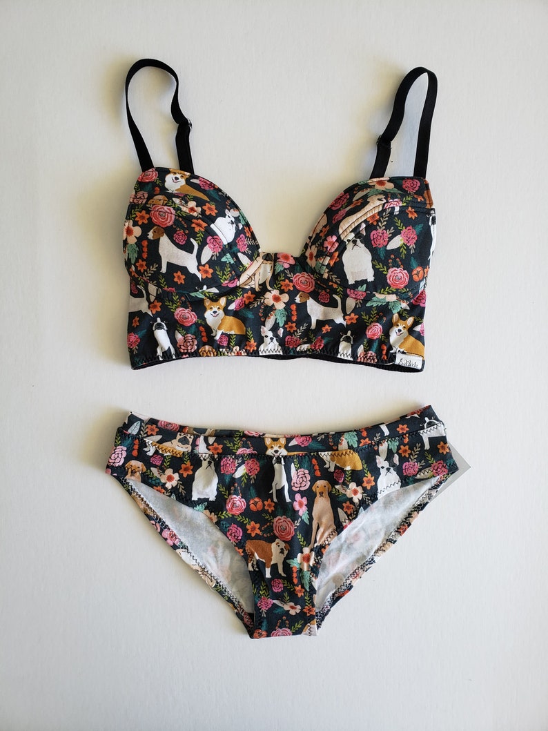 May include: Black and floral print underwear set featuring a variety of dog breeds. The bra has adjustable straps and a hook and eye fastening. The panty has a low rise and full back coverage.