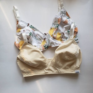 May include: Two bras, one with a floral print and the other a solid cream colour. The floral bra has a white background with yellow, orange, and green flowers and bees. The cream bra has a label that says "Edible".