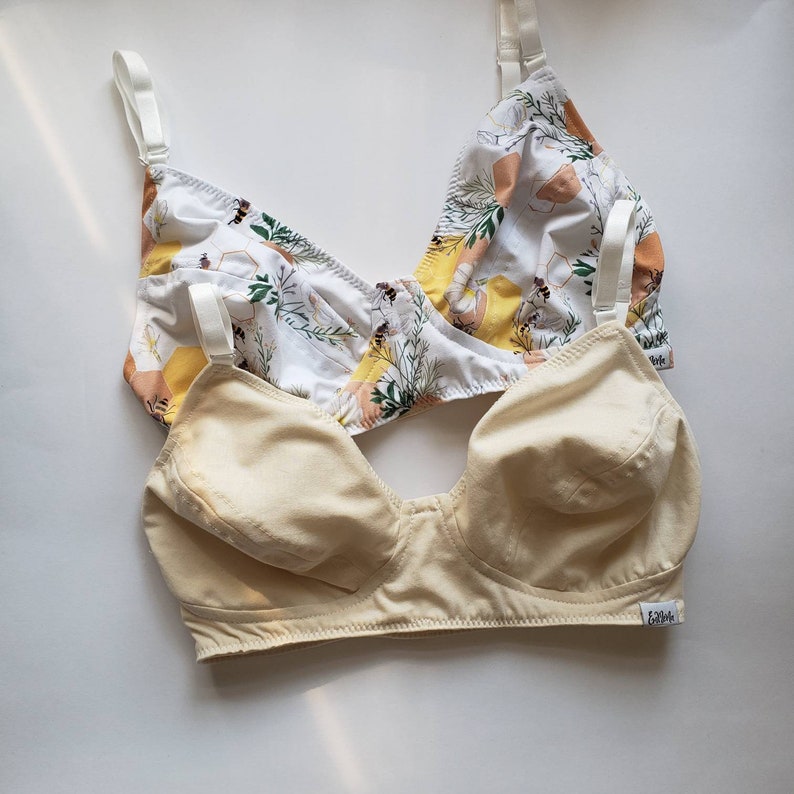 May include: Two bras, one with a floral print and the other a solid cream colour. The floral bra has a white background with yellow, orange, and green flowers and bees. The cream bra has a label that says "Edible".