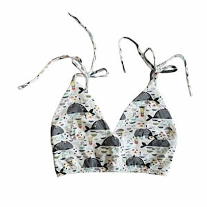 May include: A white halter top featuring a pattern of grey whales, boats, and nautical motifs. It has tie straps and a V-neck. The fabric appears soft and slightly textured. The top is designed for comfort and style.