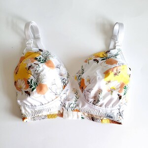 May include: White bra with adjustable straps and a floral print featuring bees and honeycomb. The bra has a yellow, orange, and white colour scheme. The brand name "Booby" is printed on the bottom band.