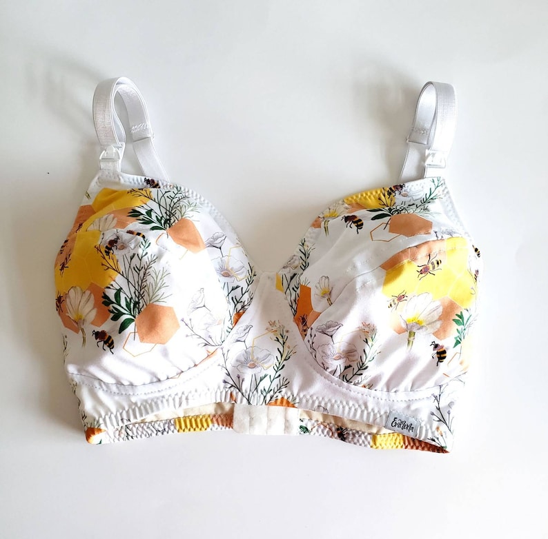 May include: White bra with adjustable straps and a floral print featuring bees and honeycomb. The bra has a yellow, orange, and white colour scheme. The brand name "Booby" is printed on the bottom band.