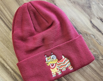 May include: A burgundy knit beanie with a folded brim. The brim features an embroidered design of a colorful lion dance figure. The hat is made of a soft, ribbed knit material.