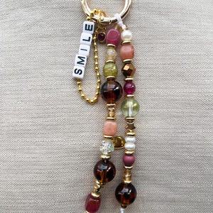 May include: A gold-colored keychain with a ring and beaded charms. The charms feature a variety of colorful beads in shades of brown, pink, green, and white. The word "SMILE" is spelled out in white letter beads.