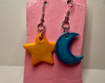 Op de afbeelding: Een paar handgemaakte oorbellen met een gele ster en een blauwe halve maan. De oorbellen hangen aan zilverkleurige haken en worden gepresenteerd op een roze achtergrond. De ster en maan zijn waarschijnlijk gemaakt van klei of een vergelijkbaar materiaal.