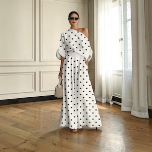 May include: A two-piece set consisting of a white, off-the-shoulder top with black polka dots and matching wide-leg trousers featuring the same black polka dot pattern. The outfit is paired with a small, textured beige handbag and dark sunglasses.