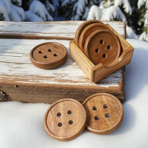 Puede incluir: Posavasos de madera con forma de botón y soporte. Los posavasos marrones, cada uno con cuatro agujeros, se asemejan a botones grandes. El juego incluye seis posavasos y un soporte de madera, ideal para proteger superficies. Se muestran sobre una superficie de madera.