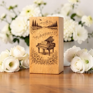 May include: A light-colored wooden box with a detailed engraving of a grand piano, musical notes, and a scenic landscape featuring mountains, trees, and a lake. Floral patterns frame the piano. The box is rectangular and stands upright.