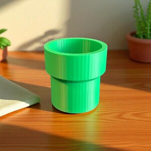 May include: A vibrant green, cylindrical container, reminiscent of a video game pipe. The container is made of a solid material and has a wide opening. It sits on a wooden surface, with a small plant visible in the background.