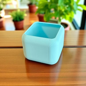 Può includere: Un vaso quadrato azzurro con angoli arrotondati. Il vaso ha un design strutturato e a coste sui lati e un interno liscio. Il vaso è su una superficie di legno. Il vaso è progettato per piccole piante.