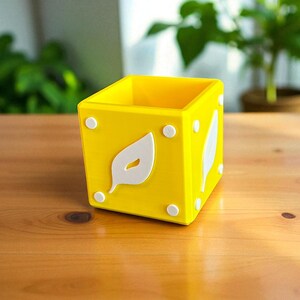 Può includere: Un vaso giallo brillante a forma di cubo con disegni di foglie bianche e accenti circolari. Il vaso è appoggiato su una superficie di legno, con piante verdi visibili sullo sfondo. È un oggetto decorativo.