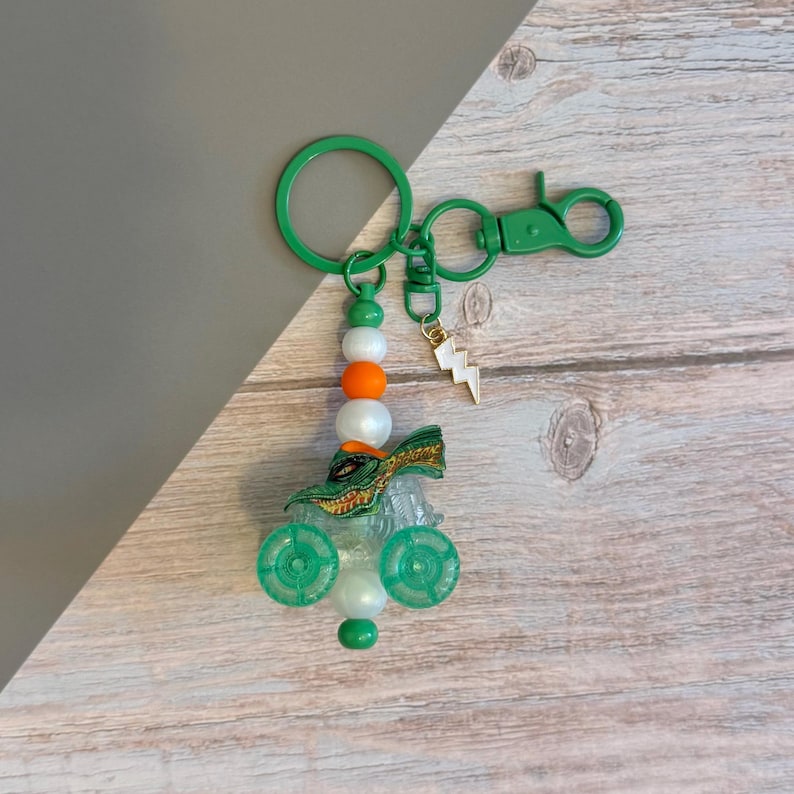 May include: A green and clear keychain featuring a small alligator figure, green wheels, and a lightning bolt charm. The keychain has a green ring and clasp, with white, orange, and green beads. The background is a mix of gray and wood.