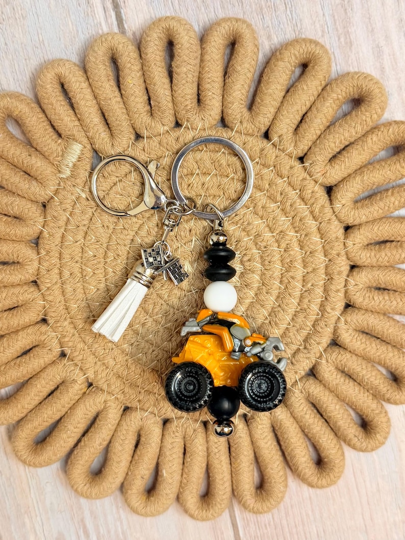 K&ouml;nnte beinhalten: Ein Schl&uuml;sselanh&auml;nger mit einem orangefarbenen ATV-Anh&auml;nger, schwarzen R&auml;dern und einem silbernen Schl&uuml;sselring. Er enth&auml;lt einen wei&szlig;en Quastenanh&auml;nger und dekorative Perlen. Der Hintergrund ist ein beigefarbener, gewebter Untersetzer.