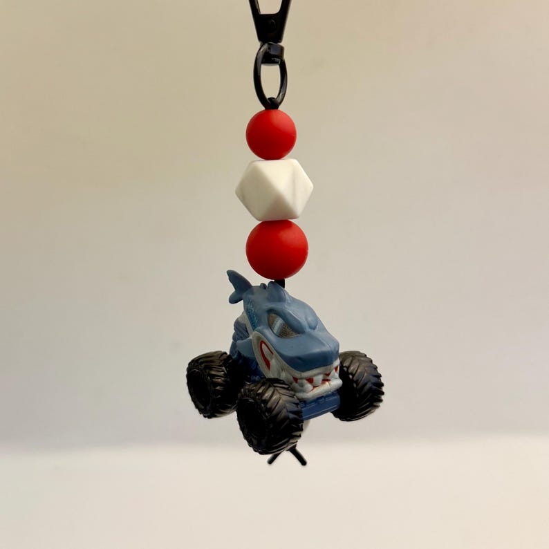 May include: A shark-themed monster truck keychain. The blue and gray truck has large black tires and a shark face design. Red and white beads are strung above the truck, attached to a black clasp.