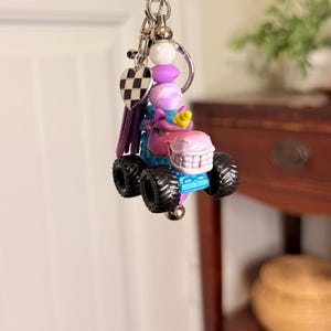 May include: A whimsical keychain featuring a monster truck design. The truck is primarily blue and pink with black wheels. The keychain includes a heart charm with a black and white checkered pattern, purple tassel, and decorative beads.