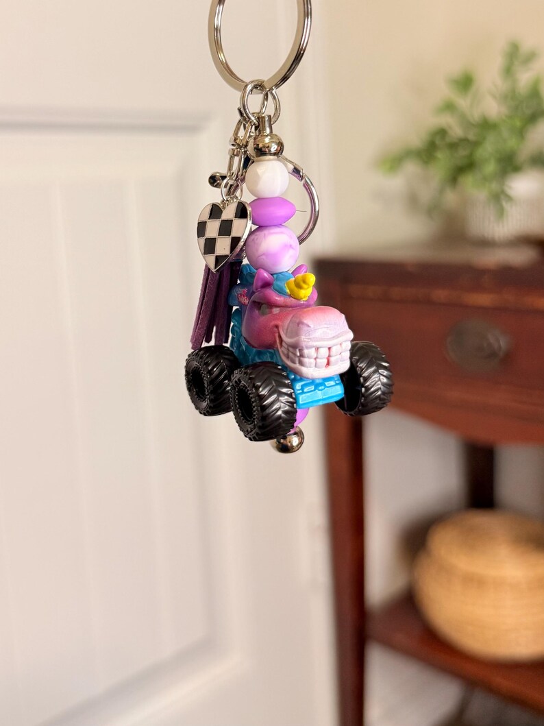 May include: A whimsical keychain featuring a monster truck design. The truck is primarily blue and pink with black wheels. The keychain includes a heart charm with a black and white checkered pattern, purple tassel, and decorative beads.