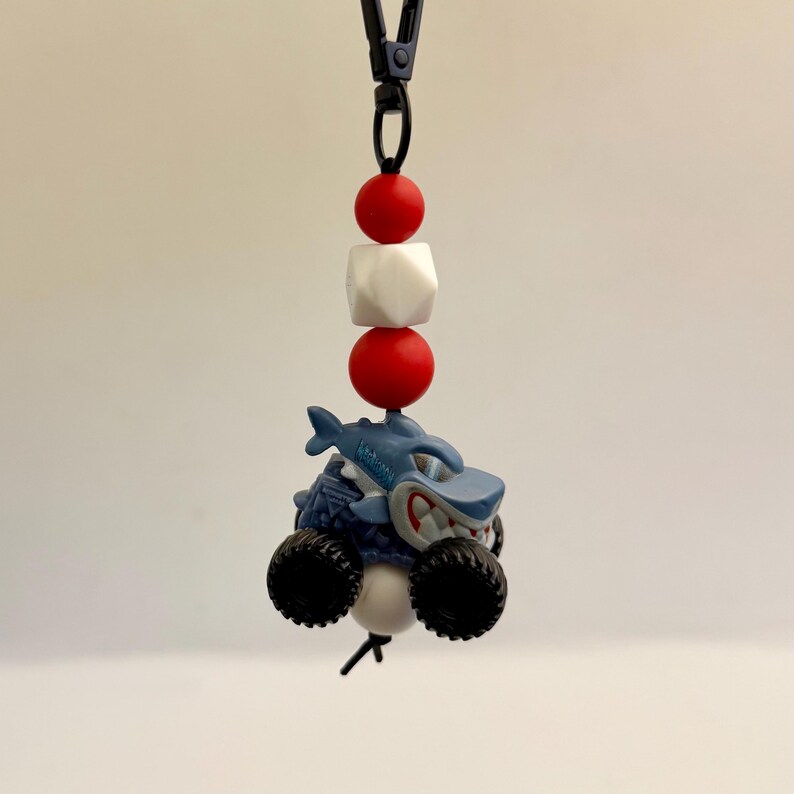 May include: A blue shark-themed monster truck toy with black wheels and a white and red bead lanyard. The shark has a menacing grin and is mounted on a blue truck body. The lanyard has red and white beads.