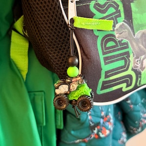 May include: A green and black monster truck charm with large tires and a green top, hanging from a backpack zipper. The charm is attached to a black string with a black and green bead.