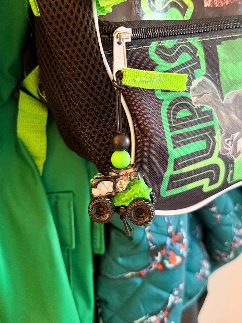 May include: A green and black monster truck charm with large tires and a green top, hanging from a backpack zipper. The charm is attached to a black string with a black and green bead.