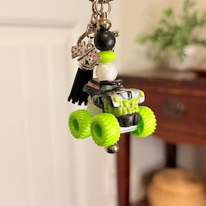 May include: A monster truck keychain with a silver keyring, black tassel, and decorative beads. The truck is black and green with neon green tires. A checkered flag charm is also attached.