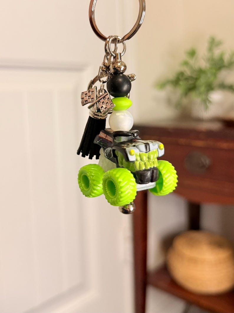 May include: A monster truck keychain with a silver keyring, black tassel, and decorative beads. The truck is black and green with neon green tires. A checkered flag charm is also attached.
