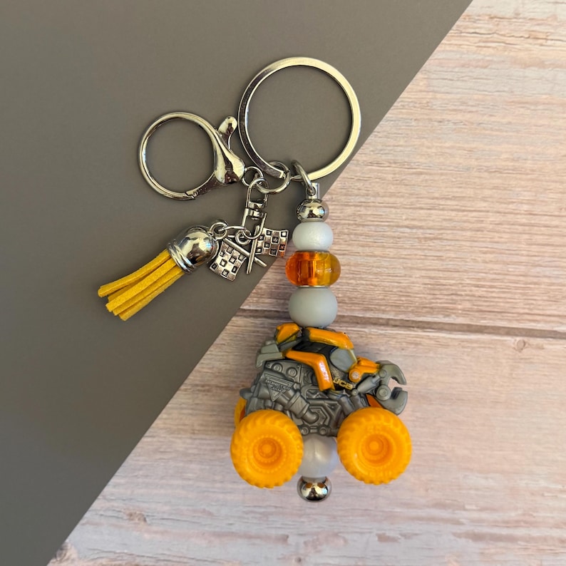 May include: A silver keyring with a yellow tassel and a monster truck charm. The truck is grey and orange with large yellow wheels. The keyring also features beads in white, grey, and orange.