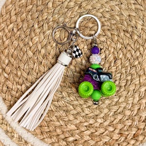 May include: A keychain featuring a miniature monster truck charm with green wheels and purple accents. The keychain includes a white tassel and a checkered flag charm, attached to a silver keyring.
