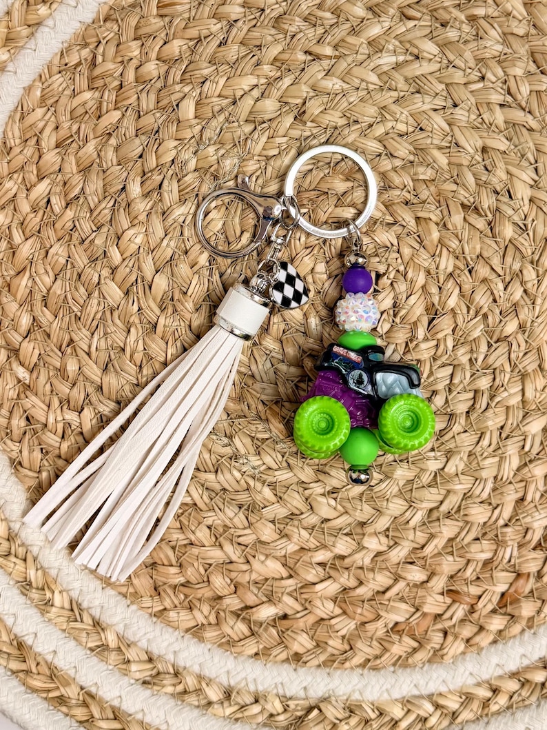 May include: A keychain featuring a miniature monster truck charm with green wheels and purple accents. The keychain includes a white tassel and a checkered flag charm, attached to a silver keyring.