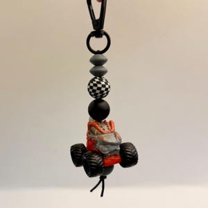 May include: A handmade keychain featuring a small monster truck toy in red and gray, with black tires. The keychain includes a series of beads in black, gray, and a checkered pattern, attached to a black clasp.