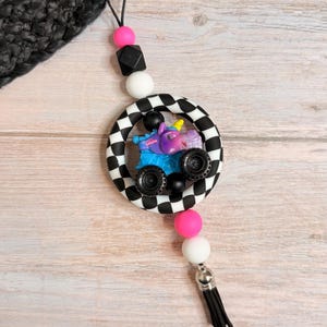 K&ouml;nnte beinhalten: Ein Autoanh&auml;nger mit einem lila Einhorn-Monster-Truck in einem schwarz-wei&szlig; karierten Kreis. Der Anh&auml;nger hat rosa, wei&szlig;e und schwarze Perlen und eine schwarze Quaste. Der Hintergrund ist ein helles Holzmaserungsmuster.