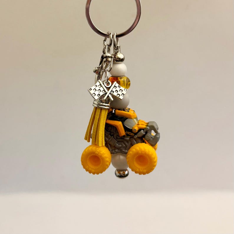 May include: A keychain with a miniature monster truck toy, featuring yellow wheels and accents. The truck is grey and orange, with a silver charm of checkered flags. The keychain also includes yellow tassels and decorative beads.