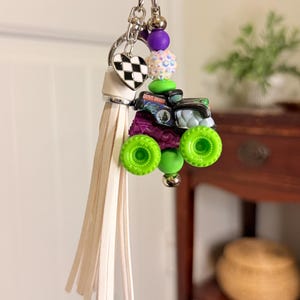 May include: A keychain featuring a miniature monster truck with green wheels and a purple body. The keychain includes a white tassel, a heart-shaped charm with a black and white checkered pattern, and various colorful beads. The words "Grave Digger" are visible on the truck.