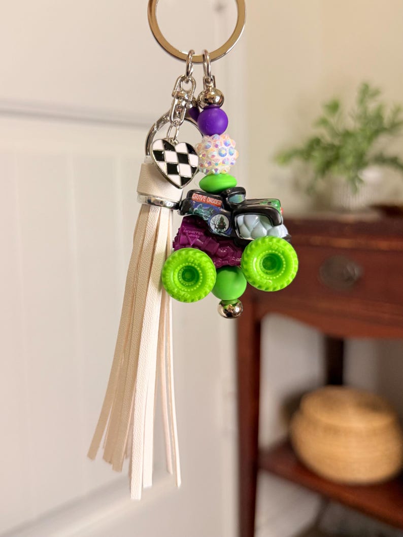 May include: A keychain featuring a miniature monster truck with green wheels and a purple body. The keychain includes a white tassel, a heart-shaped charm with a black and white checkered pattern, and various colorful beads. The words "Grave Digger" are visible on the truck.