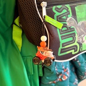 May include: A small, orange and black toy car with a tiger face is attached to a black backpack with the word "JURASSIC" printed on it. The backpack has a black mesh side pocket and green accents. A green jacket is visible in the background.
