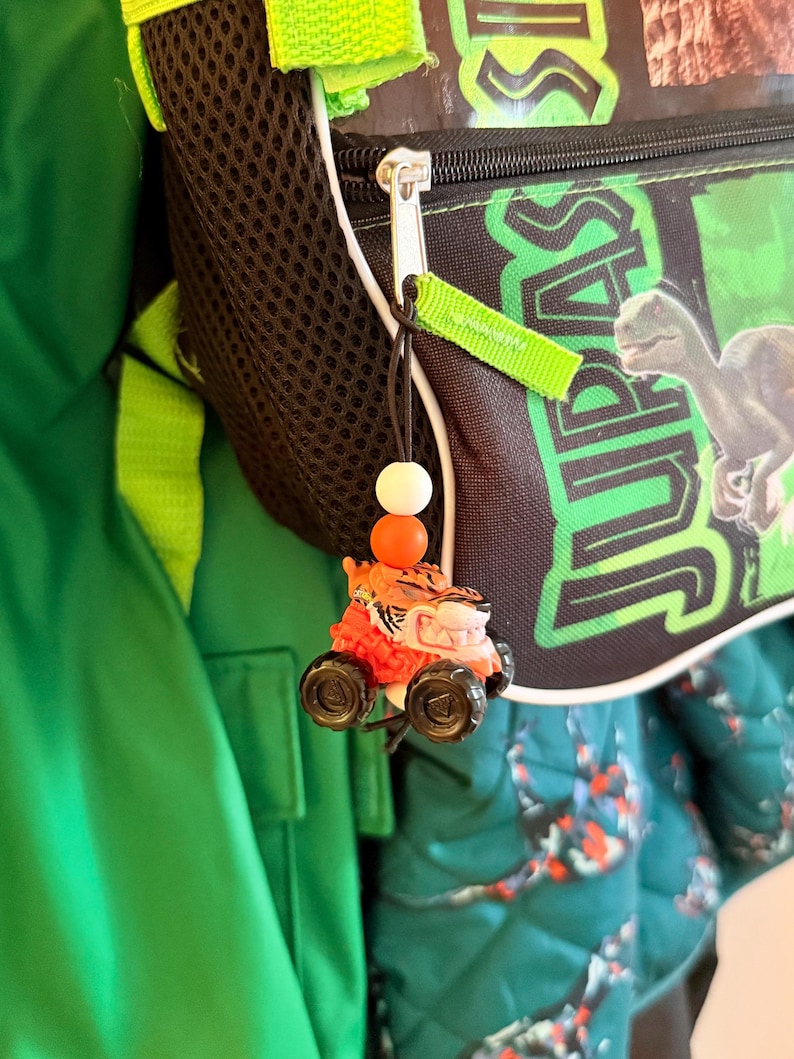 May include: A small, orange and black toy car with a tiger face is attached to a black backpack with the word "JURASSIC" printed on it. The backpack has a black mesh side pocket and green accents. A green jacket is visible in the background.