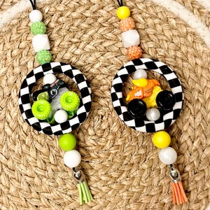May include: Two decorative lanyards featuring black and white checkered rings. The left lanyard has a green monster truck charm, lime green, white, and glittery green beads, and a lime green tassel. The right has an orange truck, yellow, white, and glittery orange beads, and an orange tassel.